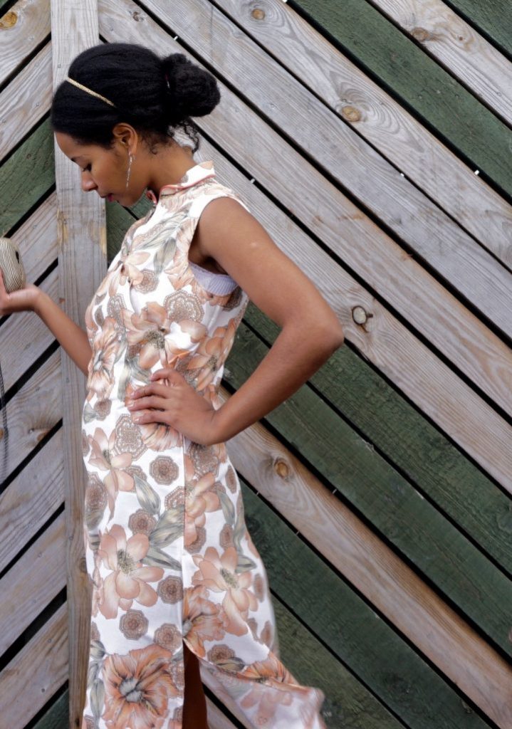 Megan is standing against a wooden fence. She is wearing her handmade qipao. The fabric has a floral print. Megan is standing sideways and she is looking down at the ground.